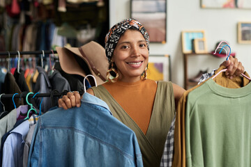 Young smiling female consumer looking at camera while visiting thrift shop and choosing secondhand...