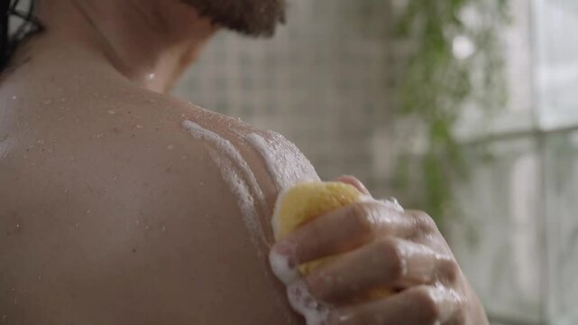 Close-up of young Caucasian man cleaning herself with soapy sponge from shoulder to neck relaxing in shower. Adult long haired male doing daily hygiene and body care alone in bathroom 