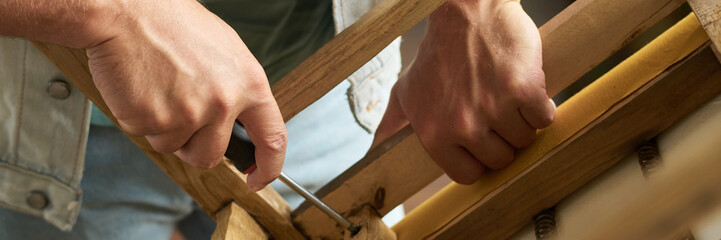 Header of hands of young unrecognizable male worker of thrift shop fixing screw in bottom part of wooden chair seat with screwdriver