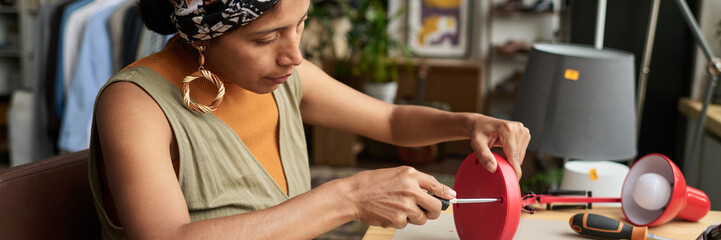 Banner of young stylish female worker of thrift shop fixing screws in electric lamp with screwdriver while sitting by workplace
