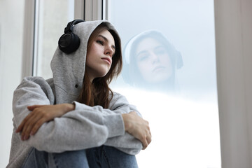 Loneliness concept. Sad teenage girl in headphones listening to music near window at home, low...