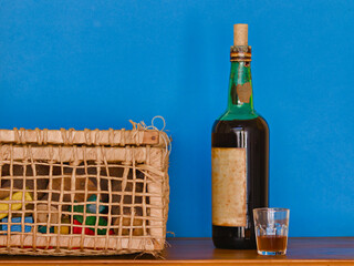 landscape image of a very old bottle of wine arranged between a glass with a shot and a wicker basket, on a wooden top, against a blue background.