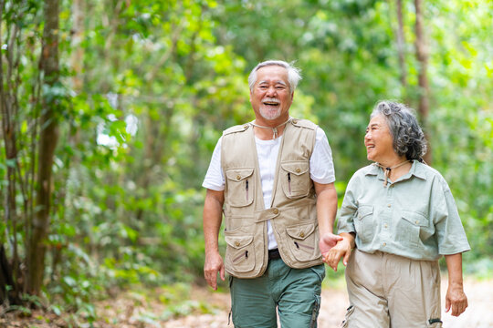 Happy Asian family senior couple walking together on tropical forest trail. Healthy elderly people enjoy and fun outdoor active lifestyle travel nature hiking and camping in jungle on summer vacation.