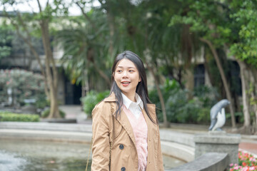 Fototapeta premium A long-haired Taiwanese woman in her 20s wearing a beige trench coat spends her winter afternoon around a fountain in a park in Xinyi District, Taipei, Taiwan, on January 30, 2024.