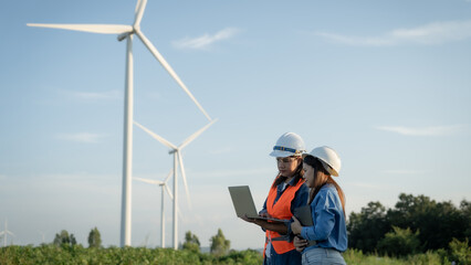 two engineers are standing in a field next to a wind farm, looking at a laptop. They are wearing...