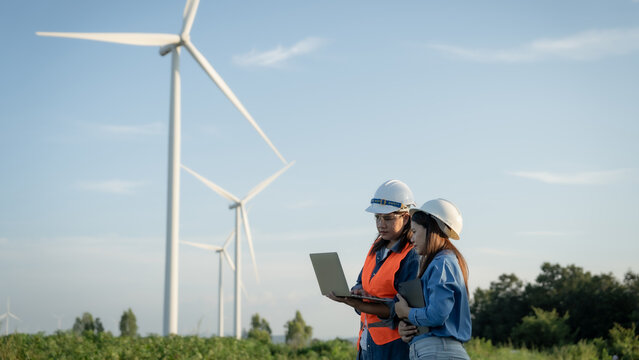two engineers are standing in a field next to a wind farm, looking at a laptop. They are wearing orange vests and hard hats, indicating that they are workers in the field. Scene is serious and focused