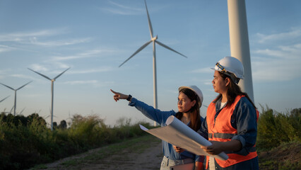 two engineers are standing in front of a wind turbine, pointing at a piece of paper. Scene is informative and professional, as the women are likely discussing the design or operation