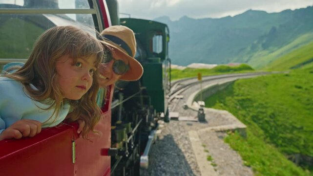 A mother and her little daughter ride in the famous red rotary steam train leaving the town of Brienz in the Swiss Alps along the alpine meadows to the top of a mountain in summer. Switzerland