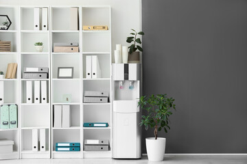 Modern water cooler with paper cups and shelf unit in office