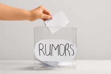 Woman putting paper into box with word RUMORS on table against light background. Gossip concept