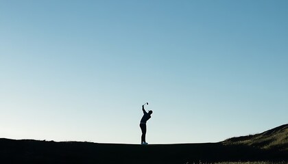 A silhouetted golfer swings on a hill against a clear blue sky at sunset.