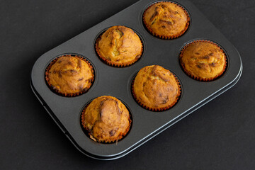 Freshly baked muffins in a muffin baking pan on dark background. Warm cupcakes in paper cups in muffin tin, baked homemade treats having a well-risen texture and slight crustiness, 