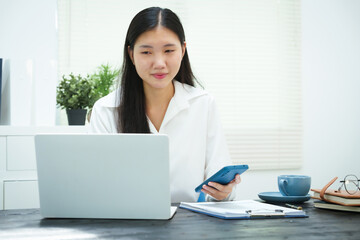 An Asian businesswoman smiles confidently while working at her desk in the office. She diligently analyzes financial data on her laptop, showcasing her expertise and commitment as an accountant
