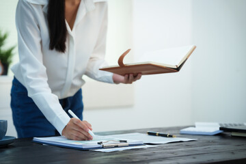 An Asian businesswoman smiles confidently while working at her desk in the office. She diligently analyzes financial data on her laptop, showcasing her expertise and commitment as an accountant