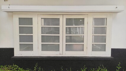 Fototapeta premium A white-framed window with six glass panes stands out against a black, textured wall base. Reflecting the outdoors, the window is bordered by greenery, adding contrast to the scene.