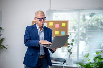 Fototapeta premium Senior businessman using laptop in modern office standing up