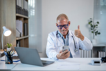 Happy doctor smiling and giving thumbs up while looking at smartphone in his office