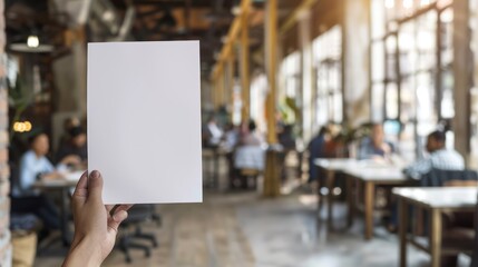 Hand Holding Blank White Paper in Front of Blurred Cafe Interior