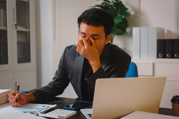 A male businessman works late at his desk, focusing on his laptop while chatting online and checking financial documents. He feels the weight of fatigue, stress, and office syndrome symptoms