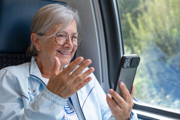 Senior woman enjoys the train ride sitting close the window talking in video call by cell phone, a...