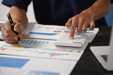 A focused businessman sits at his office desk, analyzing a paper chart while working on his laptop. He examines financial and marketing reports, concentrating on data for strategic decision-making.