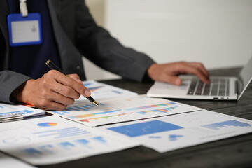 A focused businessman sits at his office desk, analyzing a paper chart while working on his laptop. He examines financial and marketing reports, concentrating on data for strategic decision-making.