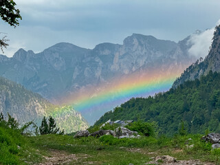 Vibrant rainbow arching over alpine village Valbona surrounded by majestic mountain massif Kolata,...