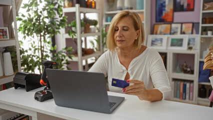 Fototapeta premium Mature woman with short blonde hair using a laptop in a decorated home office setting, holding a blue credit card, surrounded by plants and indoor decor items
