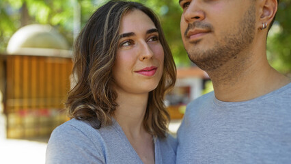 Obraz premium Woman and man standing close in a park with trees and buildings in the background, sharing an intimate moment together, both wearing gray shirts and looking content in the natural, urban setting.