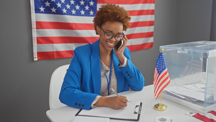 A smiling african american woman in a blue blazer taking notes in a room with an american flag