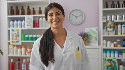 A young, attractive hispanic woman pharmacist in a lab coat smiles in a well-stocked pharmacy with shelves of products behind her.