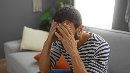 Young man with beard and striped shirt sitting on couch in living room with hands covering face looking stressed