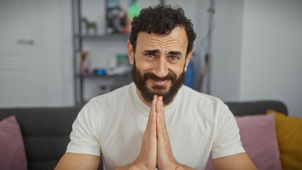 Middle-aged bearded man in white shirt gesturing prayer hands in modern living room expressing hope