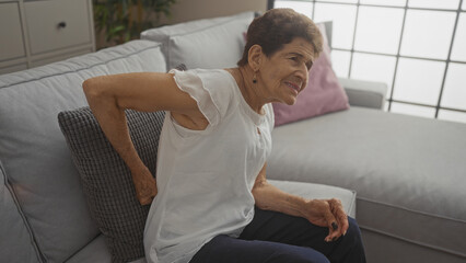 An elderly hispanic woman with short hair sitting in a living room experiencing back pain while on...