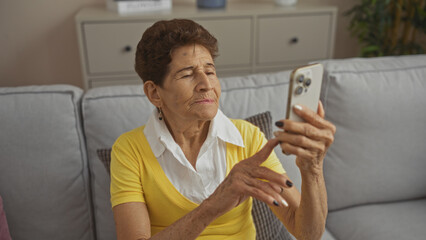 An elderly hispanic woman with short hair is seen using a smartphone while sitting on a couch in her living room, wearing a yellow top, conveying a relaxed indoor atmosphere.