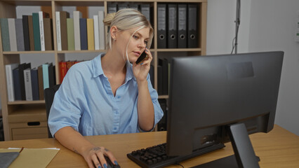 Woman talking on phone while working with a desktop computer in an office with shelves filled with...