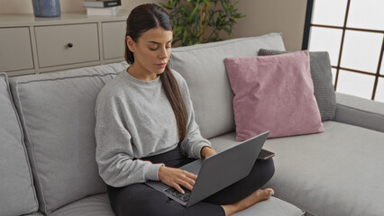 A young, attractive hispanic woman with a brunette hairstyle is working on her laptop while sitting comfortably in a modern living room of her home or apartment.