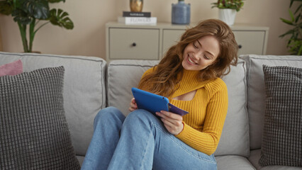 Obraz premium A young attractive blonde woman sits on a sofa at home, holding a credit card and a tablet, smiling happily in the living room.