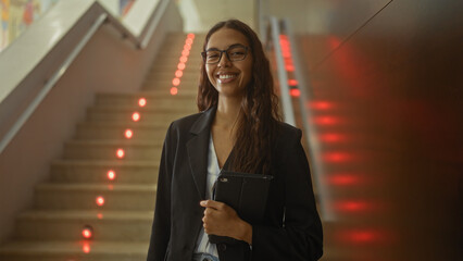 Woman smiling in a modern indoor staircase lobby holding a tablet and wearing a business suit