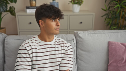 A handsome young hispanic man in a striped shirt sits on a sofa in a cozy living room with plants and modern decor.