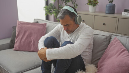 Middle-aged hispanic man sitting in a living room with headphones on, appearing pensive and sad in...