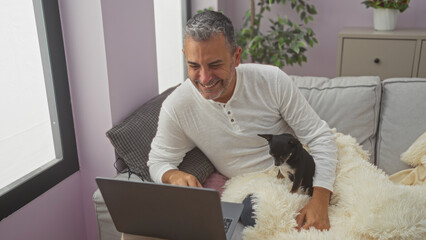 A middle-aged hispanic man smiles while sitting in his living room with his chihuahua dog, using a laptop on a cozy couch.