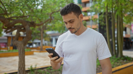 Fototapeta premium A handsome hispanic man with a beard, dressed in a white t-shirt, stands in an urban park looking at his phone on a sunny day.