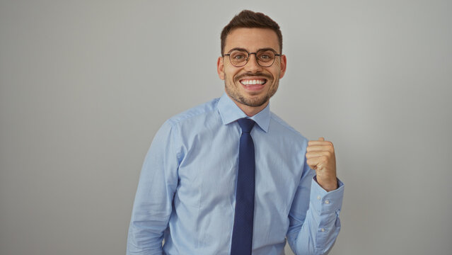 A young hispanic man with a beard and glasses in a blue shirt and tie stands isolated against a white background with a confident smile and clenched fist.