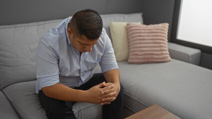 Young man sitting in a living room, looking down with hands clasped together, portraying a...