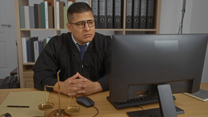 Young hispanic man wearing glasses and judge's robe sits at a desk in an office interior, focusing...