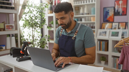 Young hispanic man working on laptop in a home decor shop, surrounded by various decorations and plants, wearing an apron with a focused expression.