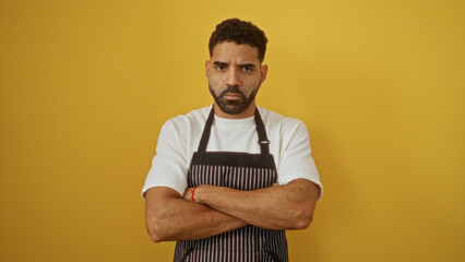 Young attractive man with beard wearing apron over isolated yellow background, looking serious with...