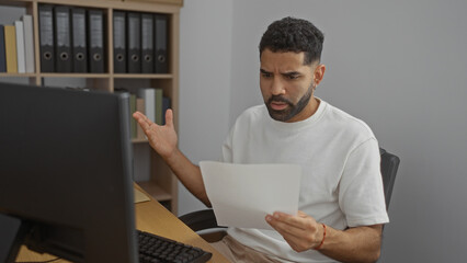 Young man looking at a document in confusion at his office desk with shelves of binders in the...