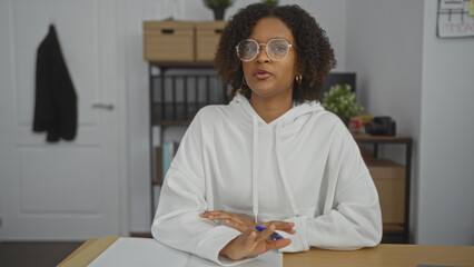 A young african american woman with curly hair and glasses is sitting at a desk in an office,...
